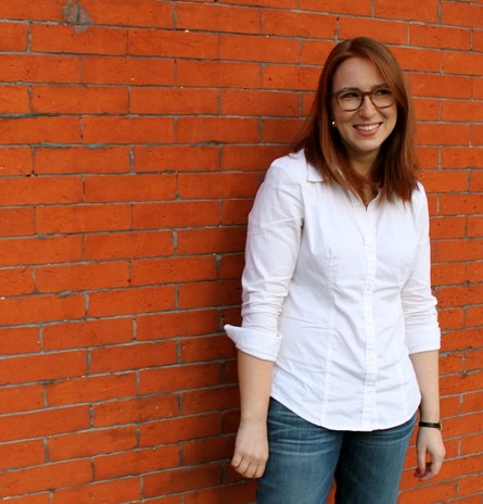 Jen, a redhead with glasses wearing a white shirt and jeans, poses smiling in front of a red brick wall.