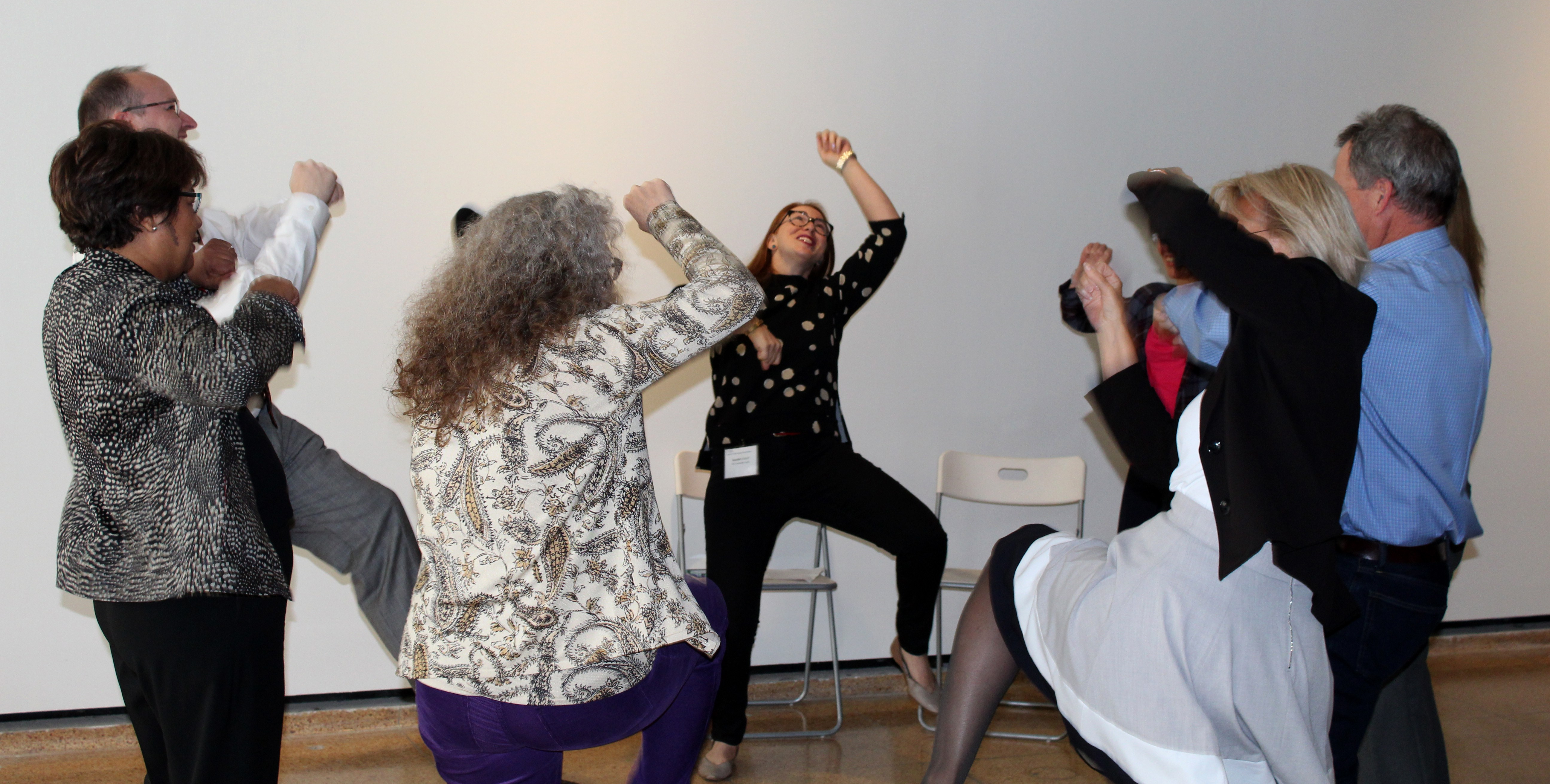 Eight people in a circle, each with one arm and one leg raised in active motion.