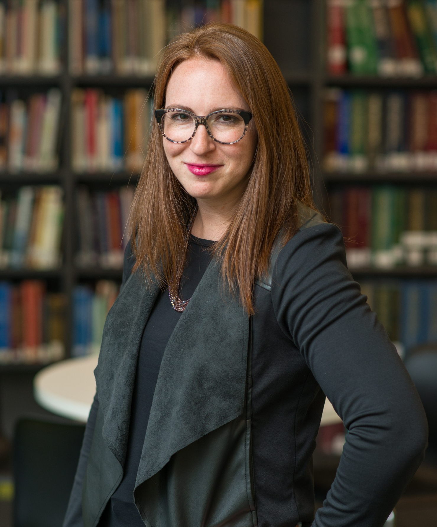 A redhead with black and white round glasses wearing a black shirt, black jacket, and a purple necklace poses in front of shelves of books. Photo courtesy of David Wolcheck.
