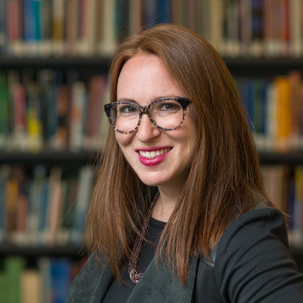 Jen Bokoff headshot. She is a redhead with glasses wearing black in front of books.