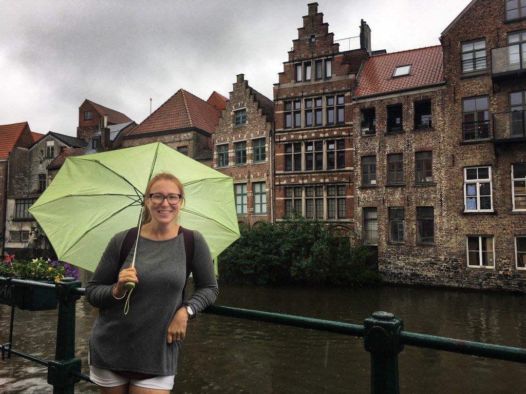 A smiling readhead with glasses and a lime green umbrella leans on a railing next to a canal.  Behind her are several brick buildings.