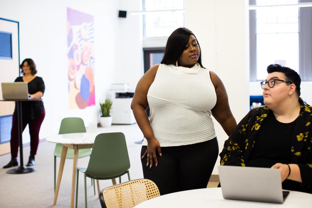 Plus-size female model stands looking at a person in glasses seated with their laptop in an office setting. Another person stands at a table with their laptop in the background.
