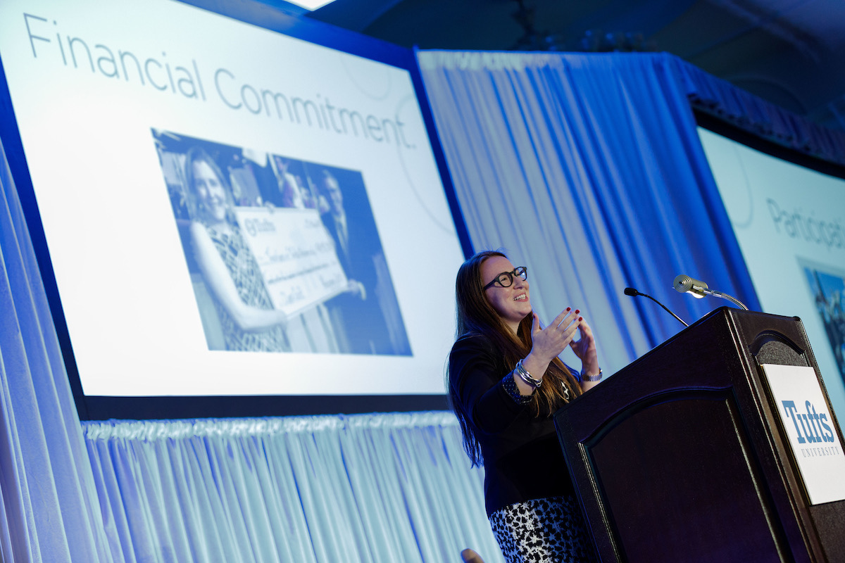 Woman with black and white outfit and speaks on large stage. The podium has Tufts University branding and behind her is a screen reading financial commitment with an image of a large check.