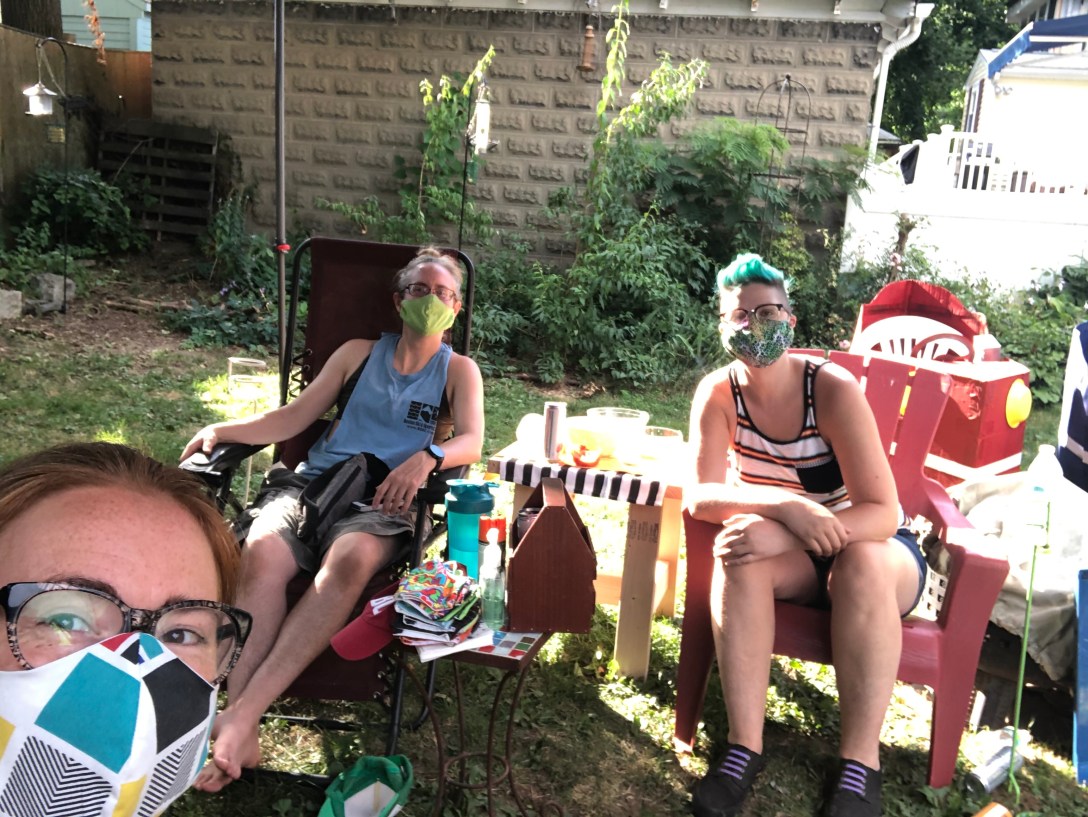 Three women sit at distance wearing masks in a yard.