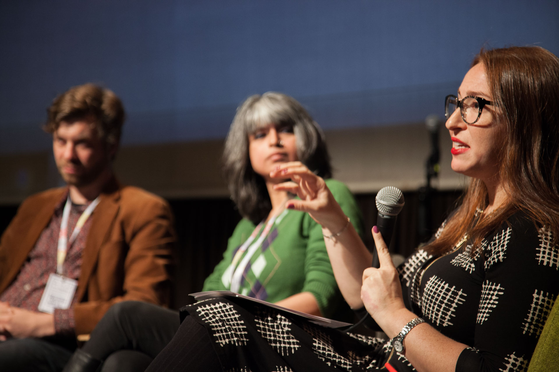 A woman in a black and white dress and glasses speaks into a microphone. A woman and a man sit next to her.
