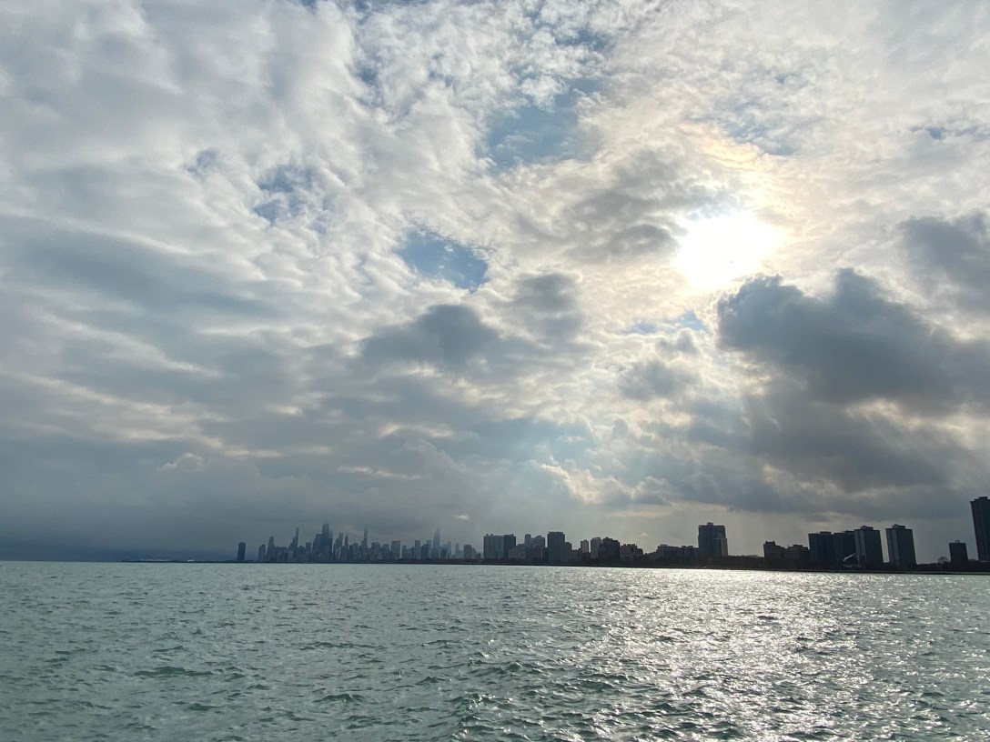 A big sky with many white clouds and the sun peaking through, creating sunbeams. On the horizon is the skyline of downtown Chicago, below which is Lake Michigan.