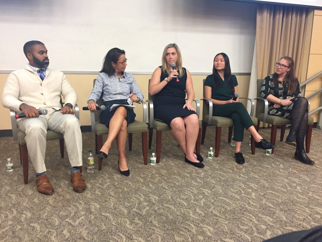 Five people sit in chairs holding microphones facing an audience. Jen is on the far right looking at the person speaking at the moment.